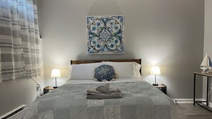 Ein ordentlich gemachtes Bett mit einem gemusterten Überwurf, zwei Nachttischlampen, einem Zierkissen und einem an der Wand hängenden Kunstwerk.