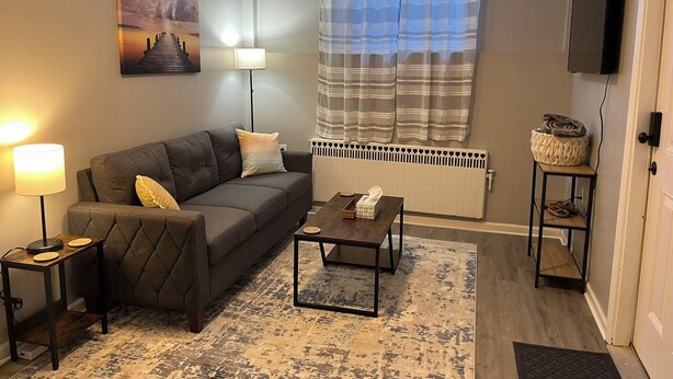 Ein Wohnzimmer mit einer Couch, zwei Beistelltischen mit Lampen, einem Couchtisch, einem Teppich, einer Heizung, einem Fernseher und einem Fenster mit Vorhängen.