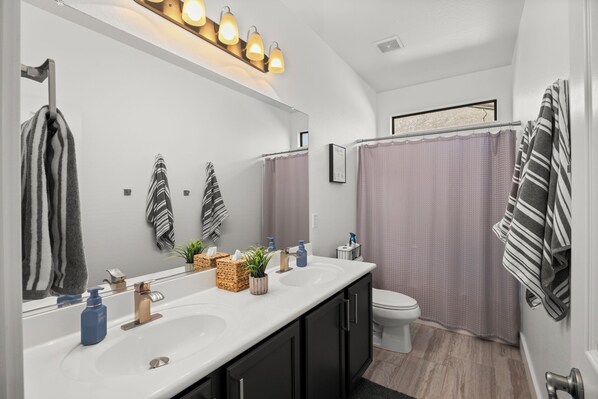 A modern bathroom with a large mirror, two sinks, a toilet, and a shower curtain. - Backyard Oasis - HTD Pool Hot Tub - Spring Training - 5BR Single Level Home (Surprise)