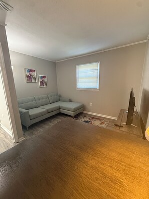 A living room with a sectional sofa, a window with blinds, a flat-screen TV on a stand, and a wooden floor. - Cozy 1-bedroom condo in Burlington, pet friendly  (Burlington)