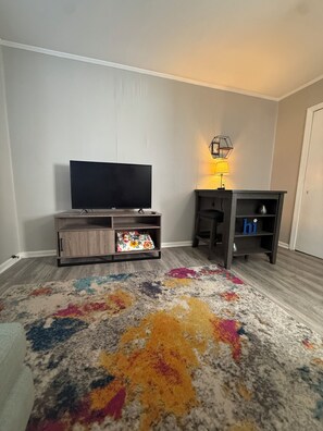 A living room with a flat-screen TV on a wooden stand, a small table with a lamp, and a colorful rug. - Cozy 1-bedroom condo in Burlington, pet friendly  (Burlington)