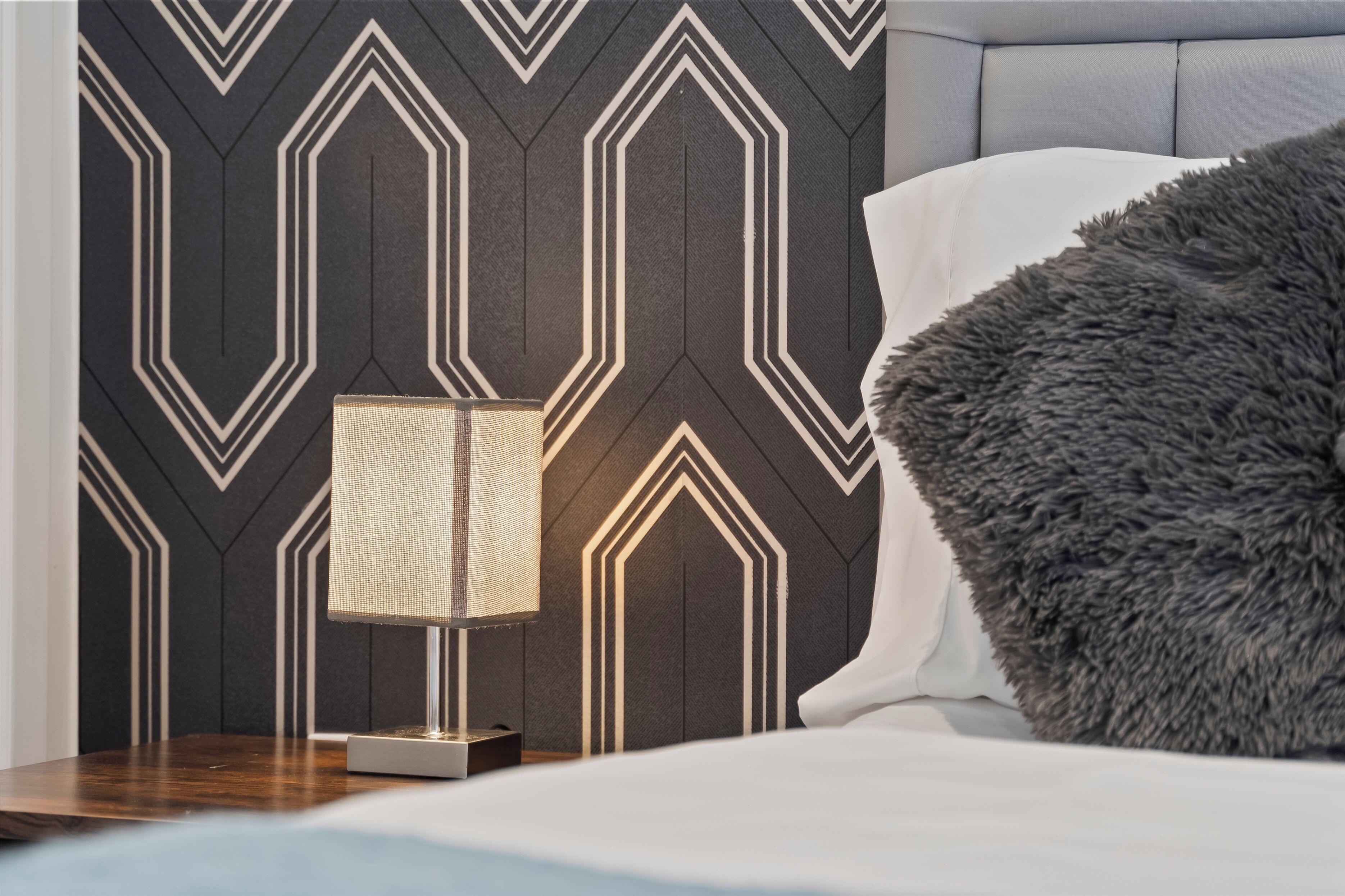 A bedroom with a geometric-patterned wall, a bedside lamp, a fluffy pillow, and a wooden nightstand.