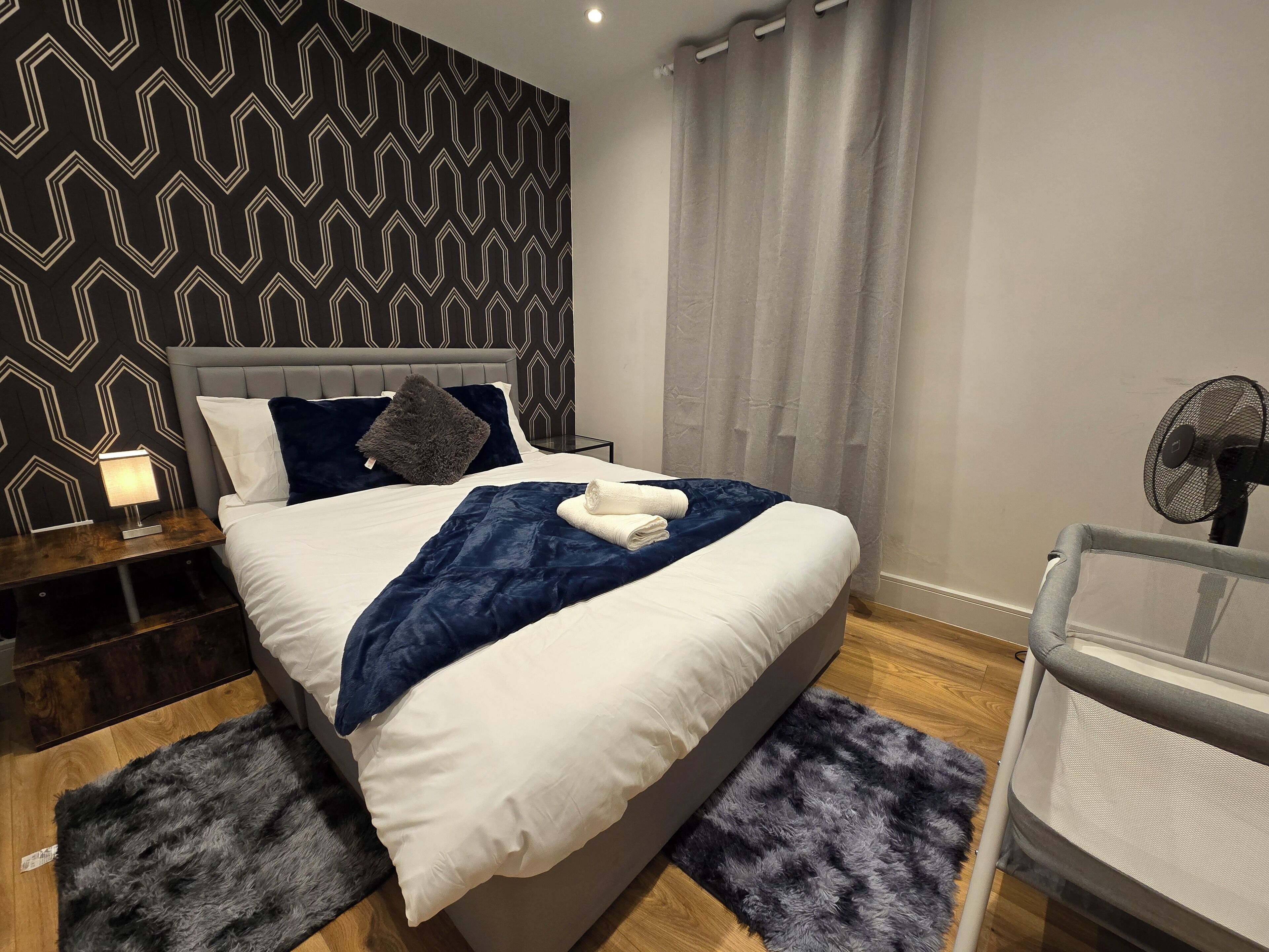 Un dormitorio moderno con una cama grande, una mesita de noche, un ventilador y una cortina gris.
