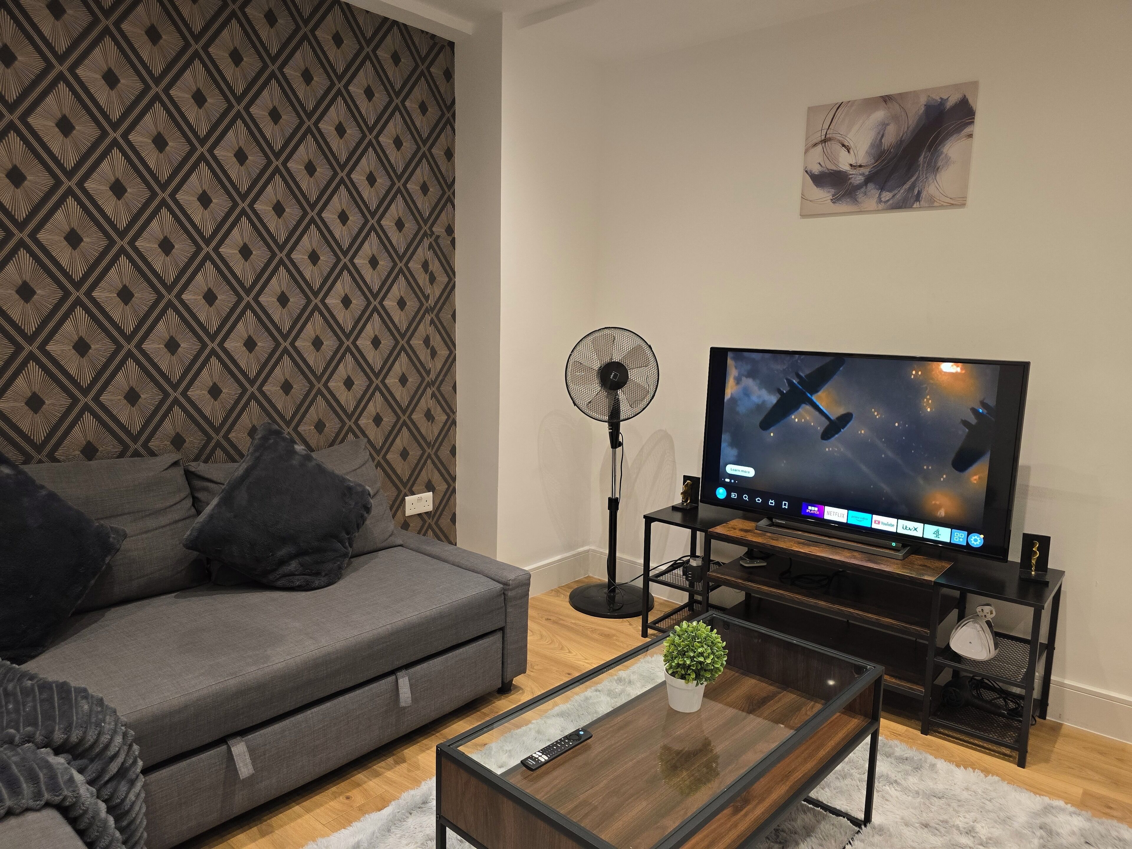 Una sala moderna con un sofá gris, una mesa de centro de madera con una planta, un televisor sobre un mueble bajo y un papel tapiz estampado.