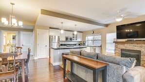 Uma cozinha moderna com uma ilha central, uma área de refeições com mesa e cadeiras de madeira, uma sala de estar com sofá e lareira, e um ventilador de teto.