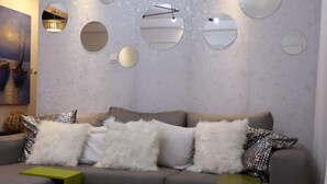 Un salon moderne avec un canapé gris, des tables d’appoint blanches et vertes, et une grande collection de miroirs accrochés au mur.