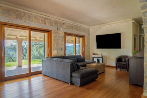 A spacious living area with a large grey sectional sofa, a small table with a lamp, and a flat-screen TV mounted on the wall.