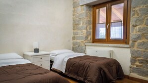 Un dormitorio con dos camas, una mesita de noche, un ventanal con vistas y paredes de piedra.