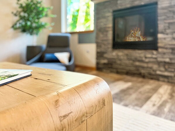 A wooden table with a book, a stone fireplace, and a chair in a room with a stone wall. - 180° mountain views • Hot Tub • 2 King Beds • Large Play Yard • AC • EV (Ashford)