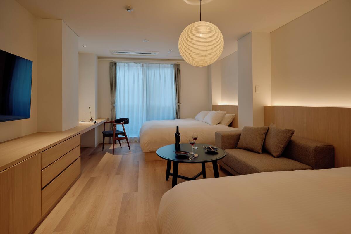 A modern hotel room with a large bed, a smaller bed, a desk with a chair, a dining table with a bottle and glass, and a large window with curtains.