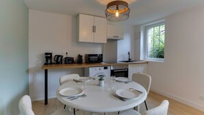 A modern kitchen with white cabinets, a dining table set for four, and a window providing natural light. - Lancelot - appt 4 pers (Bruz)