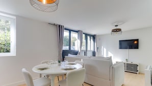 A modern living room with a dining area, a sofa, a TV, and a window with curtains. - Lancelot - appt 4 pers (Bruz)