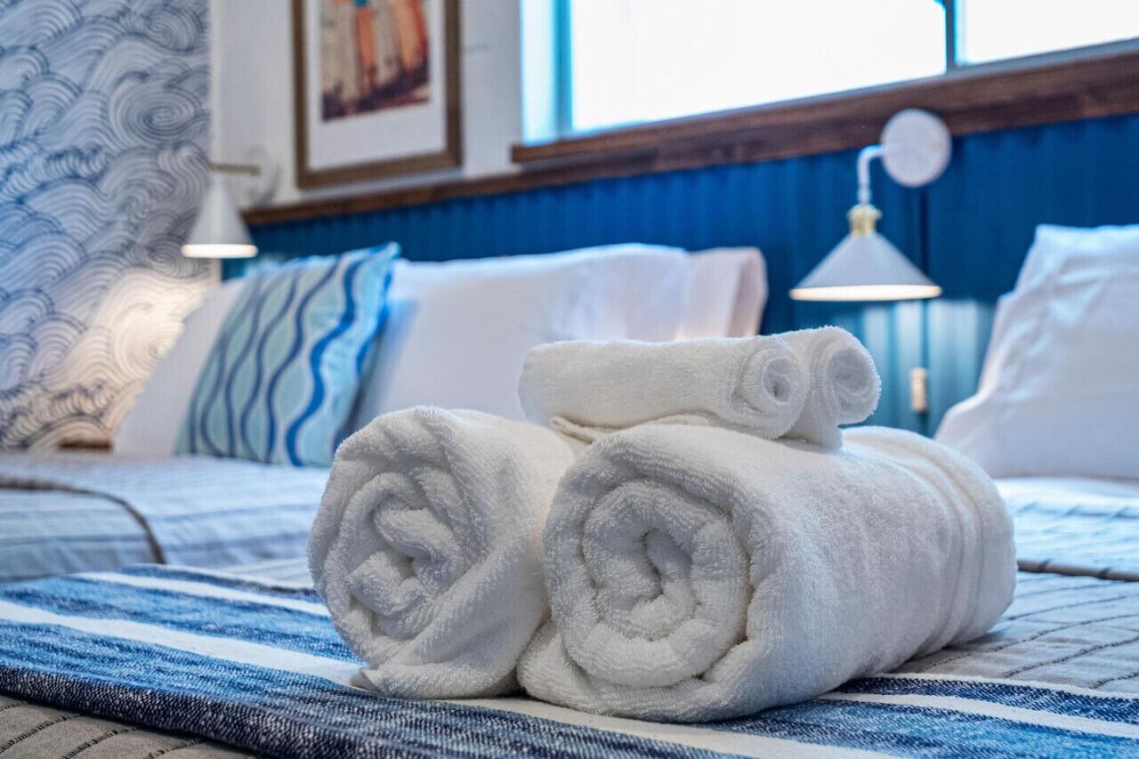 Cama bem arrumada com toalhas brancas dobradas, colcha azul e branca listrada, e parede azul com um quadro emoldurado.