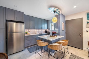 A modern kitchen with grey cabinets, a stainless steel refrigerator, a central island with bar stools, and a ceiling fan. - The SurfSide Hideout - Charming/Cozy/Central (Miami)