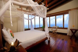 A bedroom with a large bed, wooden flooring, a view of the sea, and a balcony with a table and chair. - Gone Tropical Luxury Villa St Lucia (Delcer)