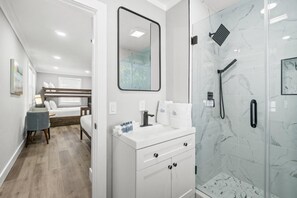A modern bathroom with a glass-enclosed shower, a vanity with a mirror, and a bed in the adjacent room. - Barefoot Beach House by PHG (Panama City Beach)