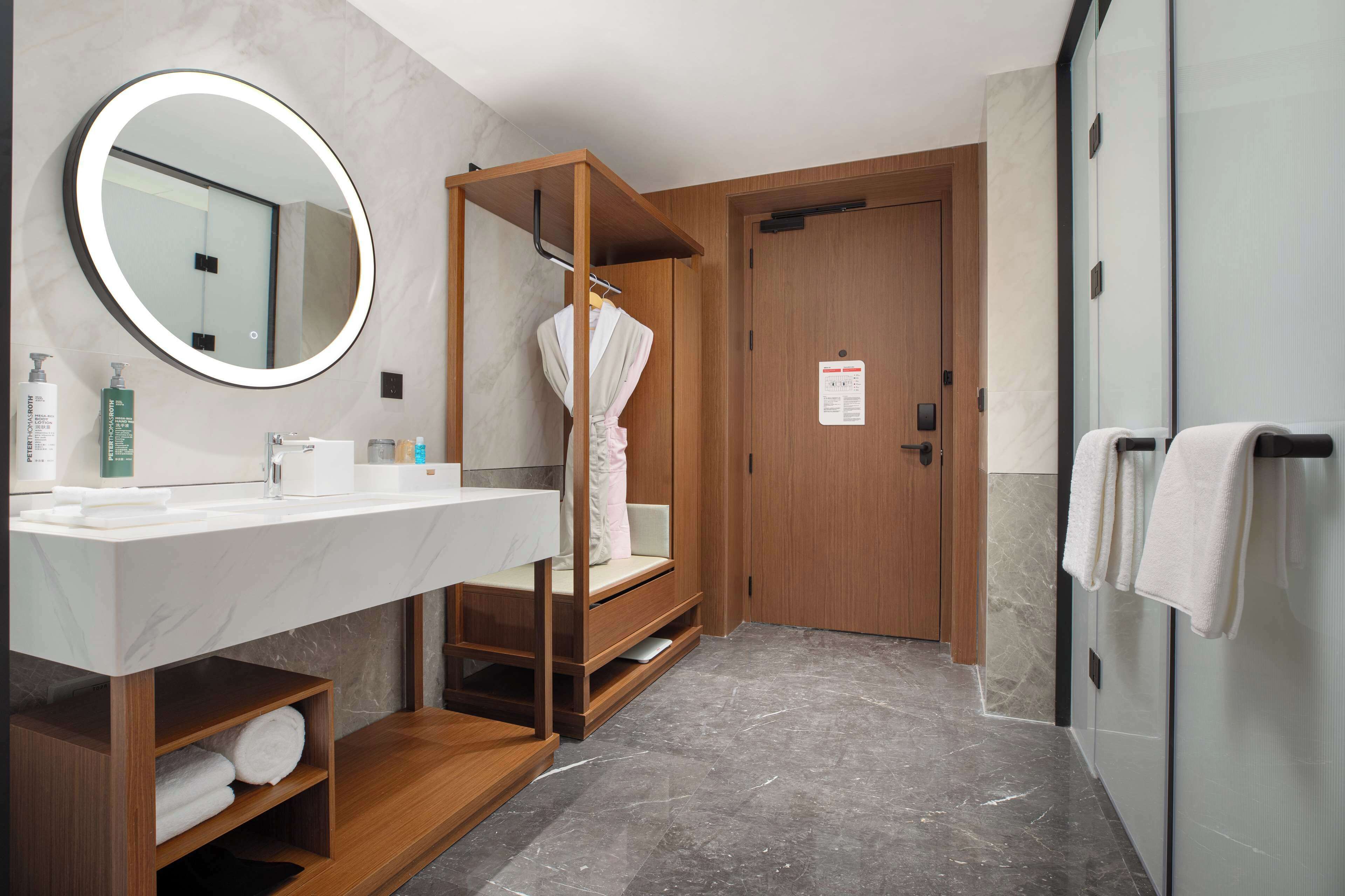 A modern bathroom with a wooden vanity, a round mirror, and a robe hanging on a rack.