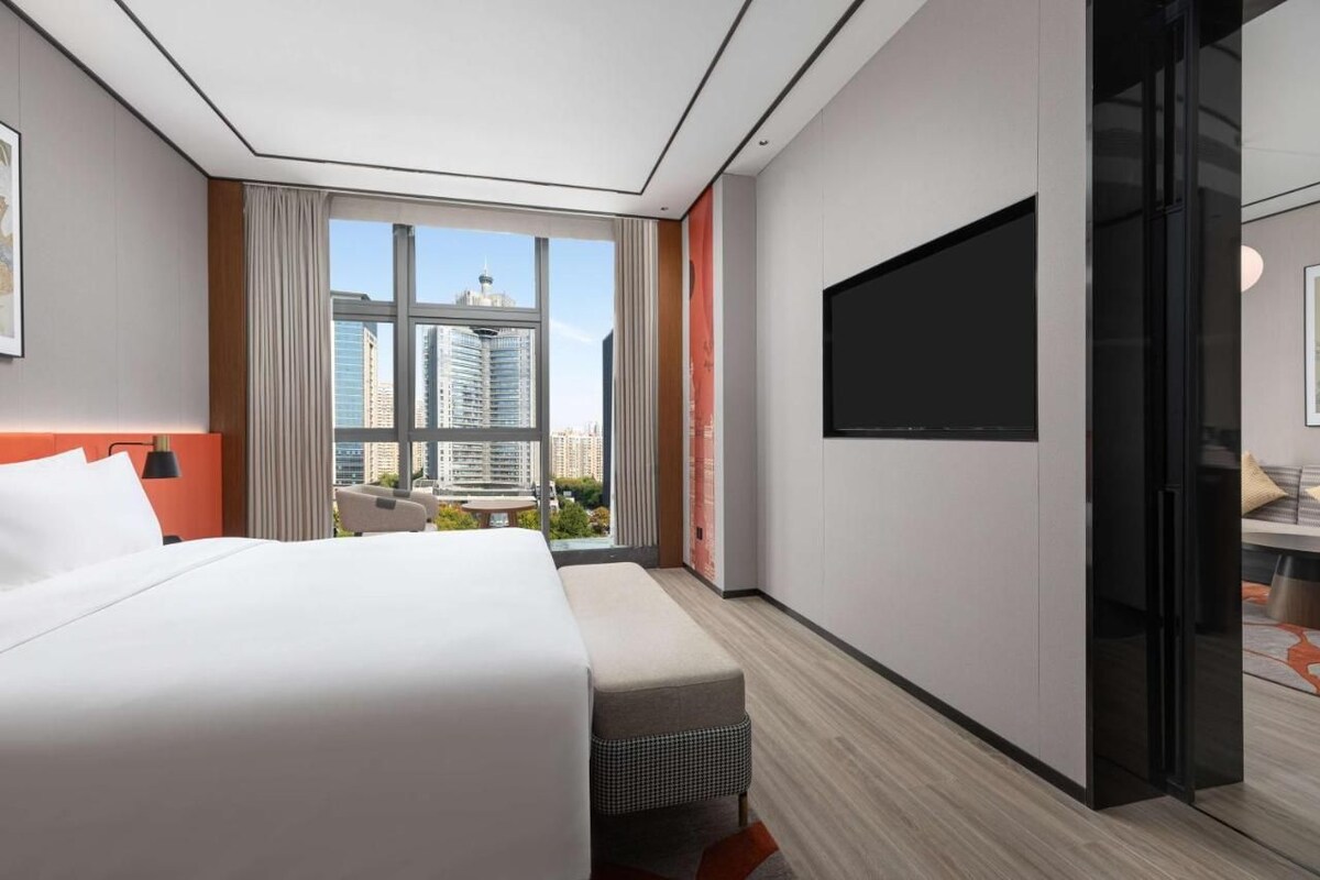Una habitación de hotel moderna con una cama grande, un televisor de pantalla plana, una zona de estar con vistas a la ciudad y un ventanal amplio.
