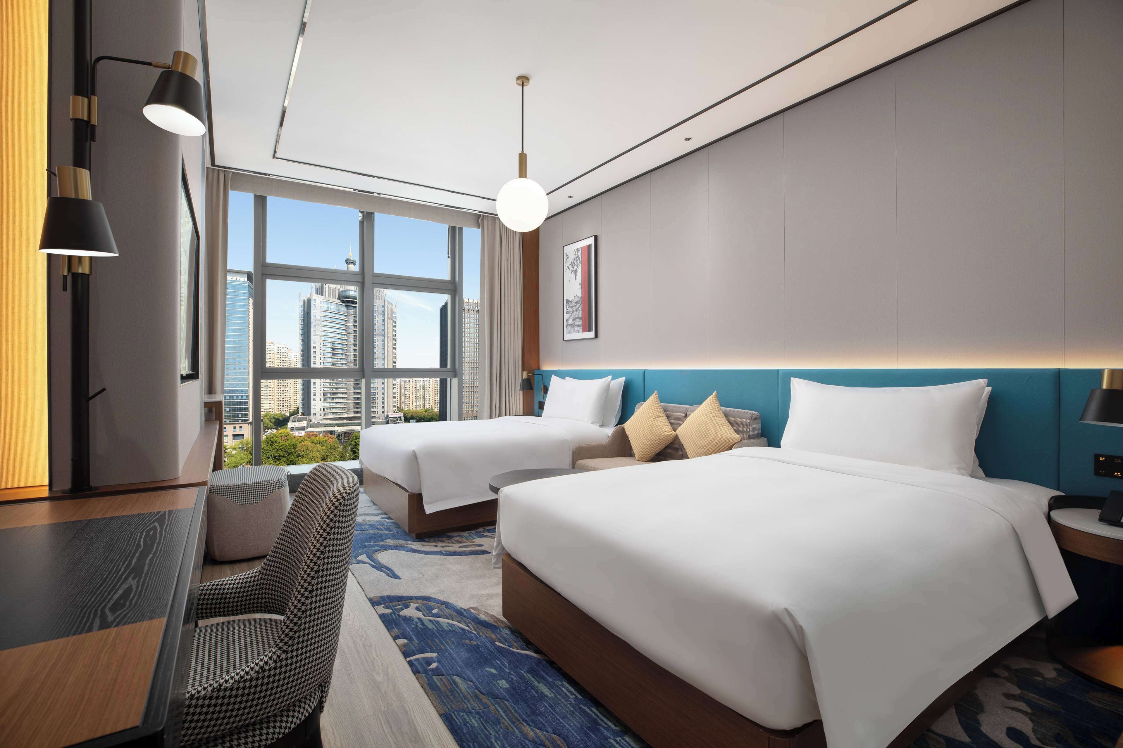 A hotel room with two beds, a desk, and a view of the cityscape.