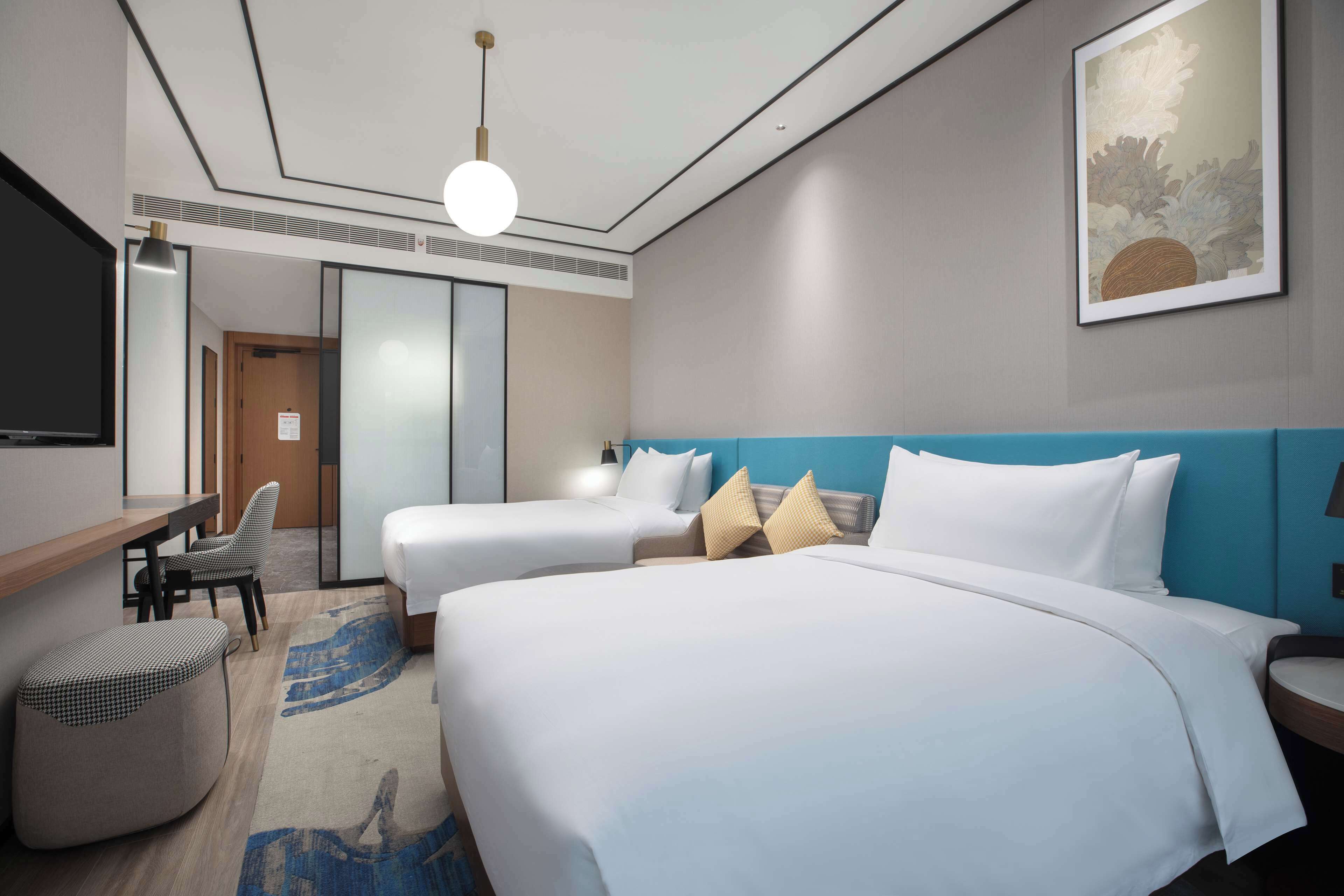 A hotel room with two beds, a desk, a chair, a large painting, and a blue accent wall.