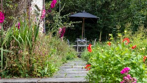 A stone pathway leading to a garden area with a metal table and chairs, surrounded by lush greenery and flowering plants.