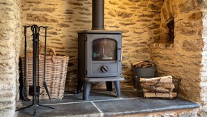 A stone-walled room with a wood-burning stove, a wicker basket, and a metal basket with logs.