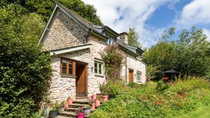 A stone house with a wooden door and windows, surrounded by lush greenery and a garden with potted plants.