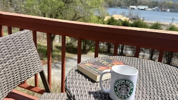 Ein Korbtisch mit einer weißen Tasse und einem Buch darauf, auf einem Balkon mit Blick auf einen See und Bäume.