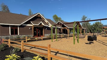 Ein Gebäude mit braunem Dach und Holzelementen, ein Spielplatz mit Schaukeln und Rutschen sowie ein Holzzaun im Vordergrund.