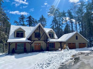A large, multi-story log cabin with a steeply pitched roof, surrounded by a snowy landscape and tall pine trees. - Bolton Bear House|Dock Slip|Hot Tub|12 Beds|Pet Friendly| (Bolton Landing)