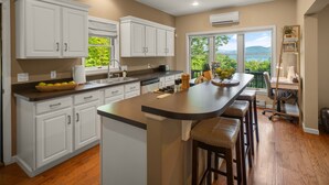 A kitchen with white cabinets, a central island with stools, a window with a view of greenery, and a small office area with a desk and chair. - Lakeview Lodge|Hot Tub|9 Beds|Pet Friendly|Dock Slip|Lake Views| (Bolton Landing)