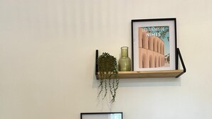 Deux étagères en bois adossées à un mur blanc, chacune portant un tableau encadré et une petite plante.