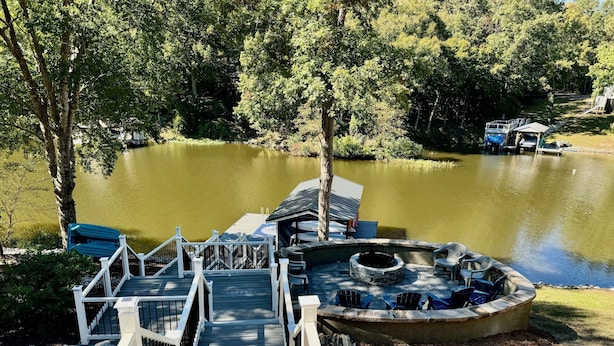 Un muelle de madera que lleva a un barco atracado, un pozo de fuego y una zona verde con sillas.