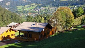 Eine Berglodge mit einer großen Holzhütte, einer Veranda und Blick auf ein Dorf sowie bewaldete Berge im Hintergrund.