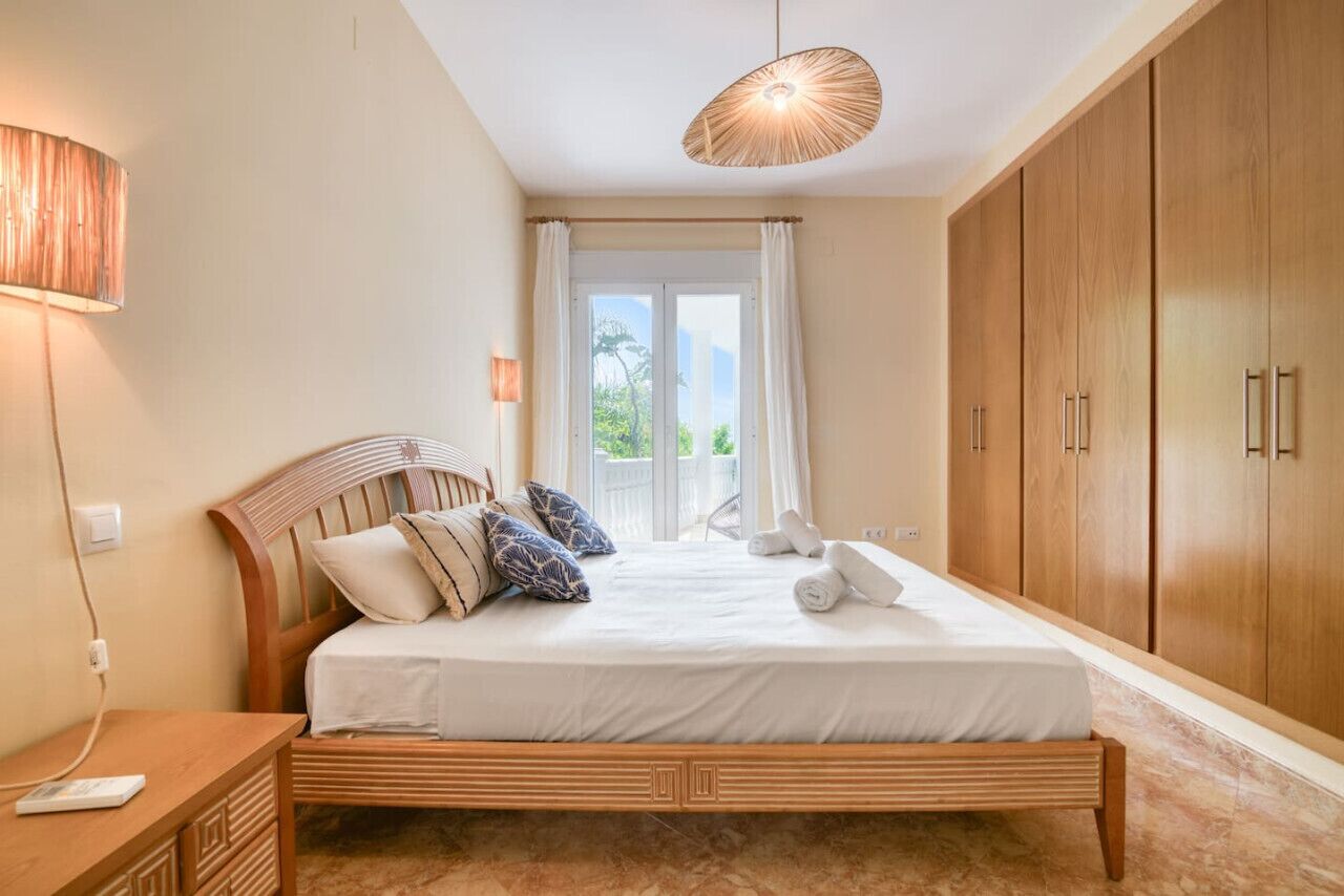 A bedroom with a wooden bed, a nightstand, a lamp, a wardrobe, and a view through a window.
