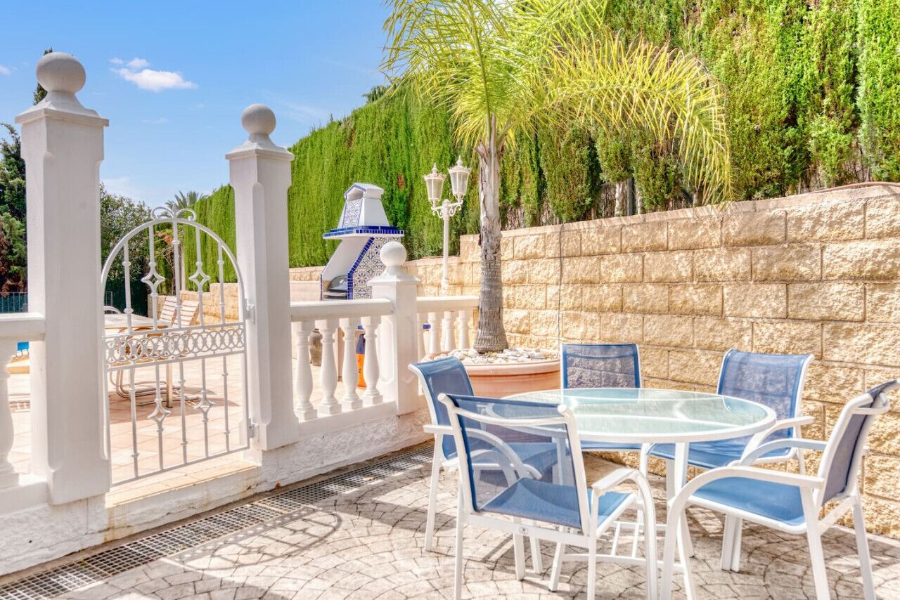 Une terrasse extérieure avec une table en verre et des chaises blanches, entourée d’une clôture blanche et d’un mur en pierre.