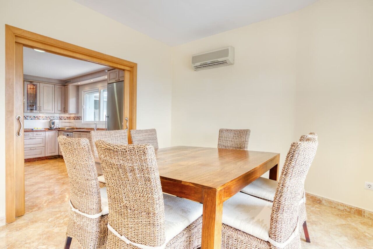 A dining area with a wooden table and wicker chairs, a kitchen in the background, and an air conditioning unit on the wall.