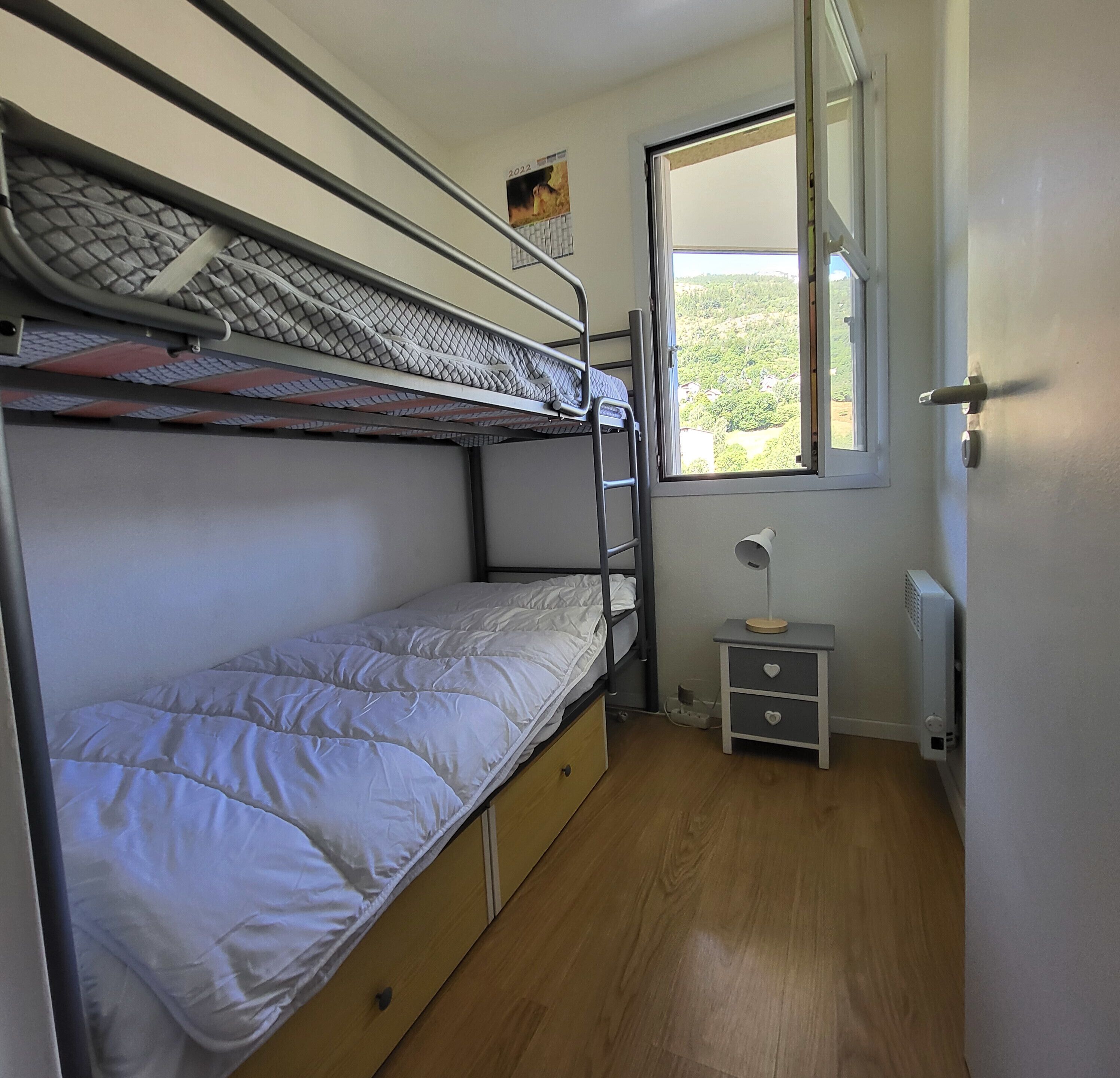 Une chambre avec un lit superposé, une fenêtre, une petite table de chevet et un radiateur.
