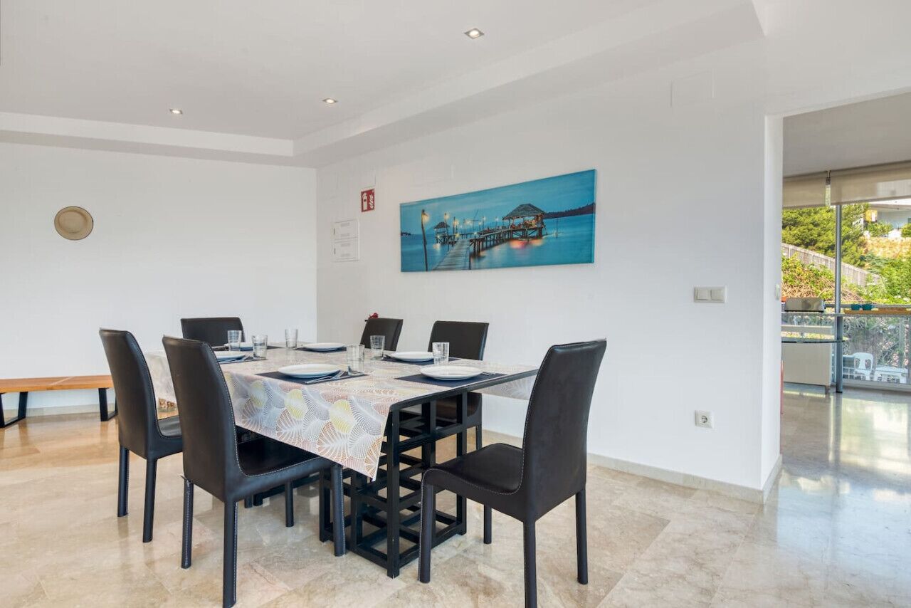 Un coin repas avec une table dressée pour six personnes, entourée de chaises noires, un tableau au mur et un banc à l’arrière-plan.