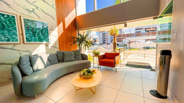A modern living room with a curved grey sofa, a small round table with a bowl of flowers, a red armchair, and a large window offering a view of the cityscape. - Branco Haus Seaview By Living (João Pessoa)