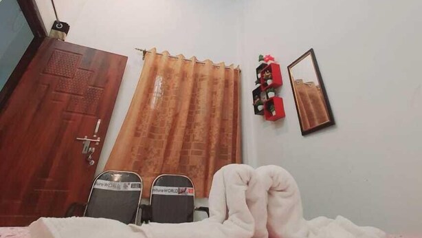 A bedroom with a ceiling fan, an air conditioner, a wooden door, a window with curtains, a bed with a pink blanket, a bedside table, and a mirror on the wall. - Kunal Guest House  (Varanasi)