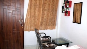 A room with a wooden door, a window with a patterned curtain, a small table with a glass top, a chair, and a shelf with decorative items. - Kunal Guest House  (Varanasi)