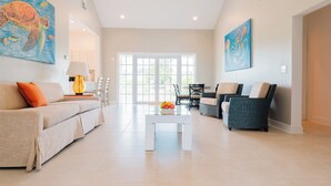 A spacious living room with a beige sofa, a white coffee table with fruit, and a large window with natural light. - Three Bedroom Superior Apartment (Jolly Harbour)