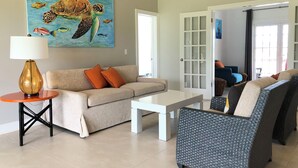 A living room with a beige sofa, orange cushions, a white coffee table, a lamp, and a painting of a sea turtle. - Three Bedroom Superior Apartment (Jolly Harbour)