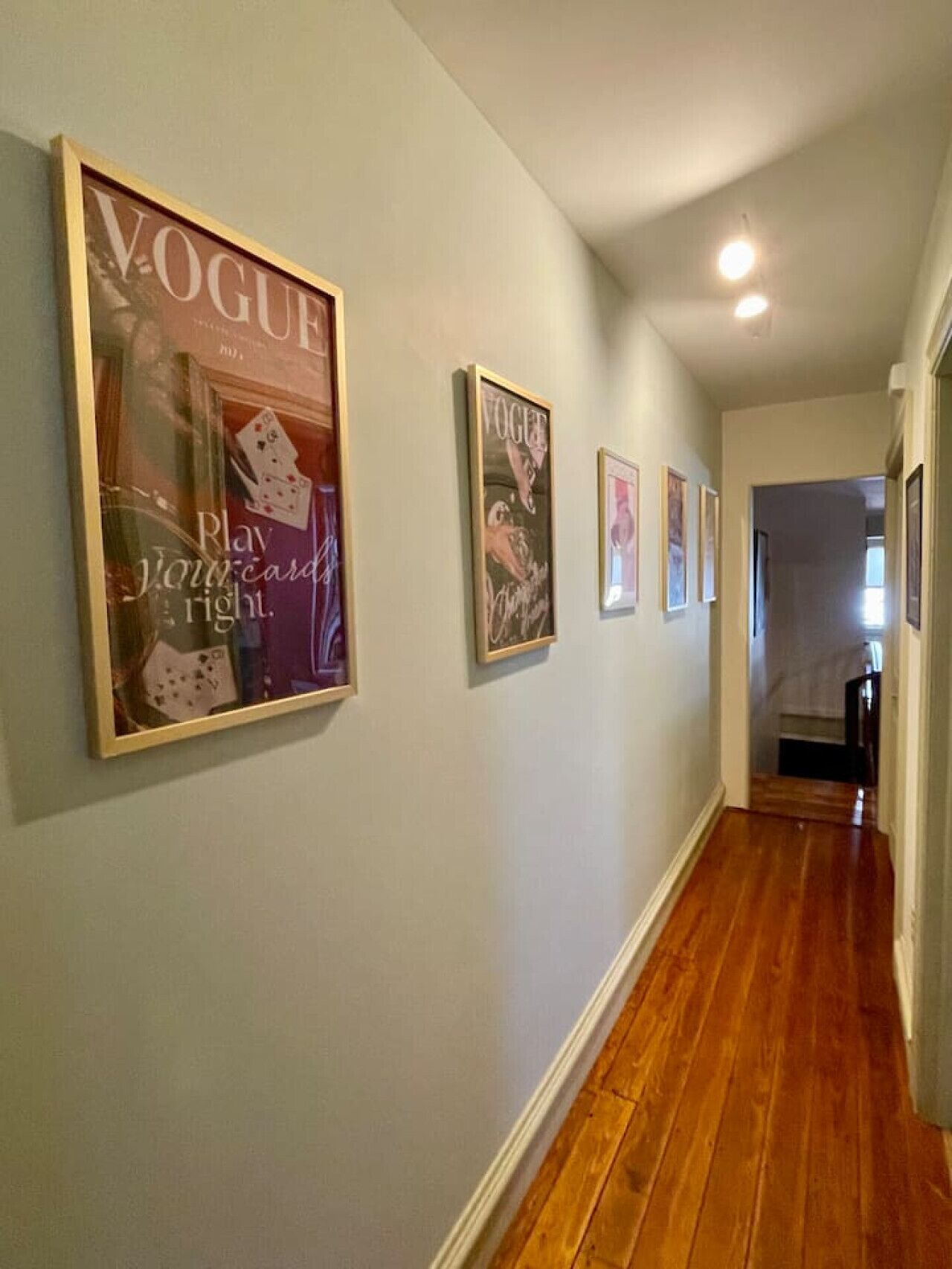 Ein Flur mit Holzdielen und hellen Wänden. An der Wand hängen drei gerahmte Poster, darunter ein Cover des Magazins Vogue.
