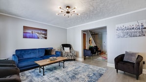 Ett modernt vardagsrum med en blå soffa, en mönstrad fåtölj och ett soffbord i trä. En trappa leder till ett annat rum, och en målning som föreställer en stadsbild hänger på väggen.