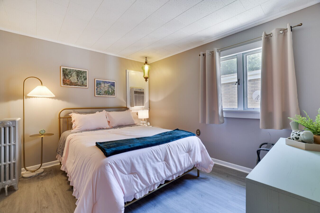 A bedroom with a bed, bedside lamps, a radiator, a window with curtains, and wall art.