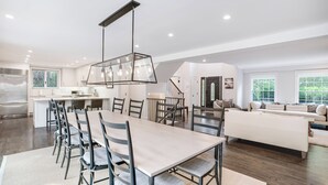 A spacious kitchen with a central island, a dining area with a long table and chairs, and a living area with a sofa and a large window. - Expansive Modern Sag Harbor Home with Private Heated Pool, Perfect for Families (Sag Harbor)
