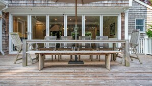 An outdoor wooden deck with a table and chairs, a covered seating area, and a view of the house through large glass doors. - Expansive Modern Sag Harbor Home with Private Heated Pool, Perfect for Families (Sag Harbor)