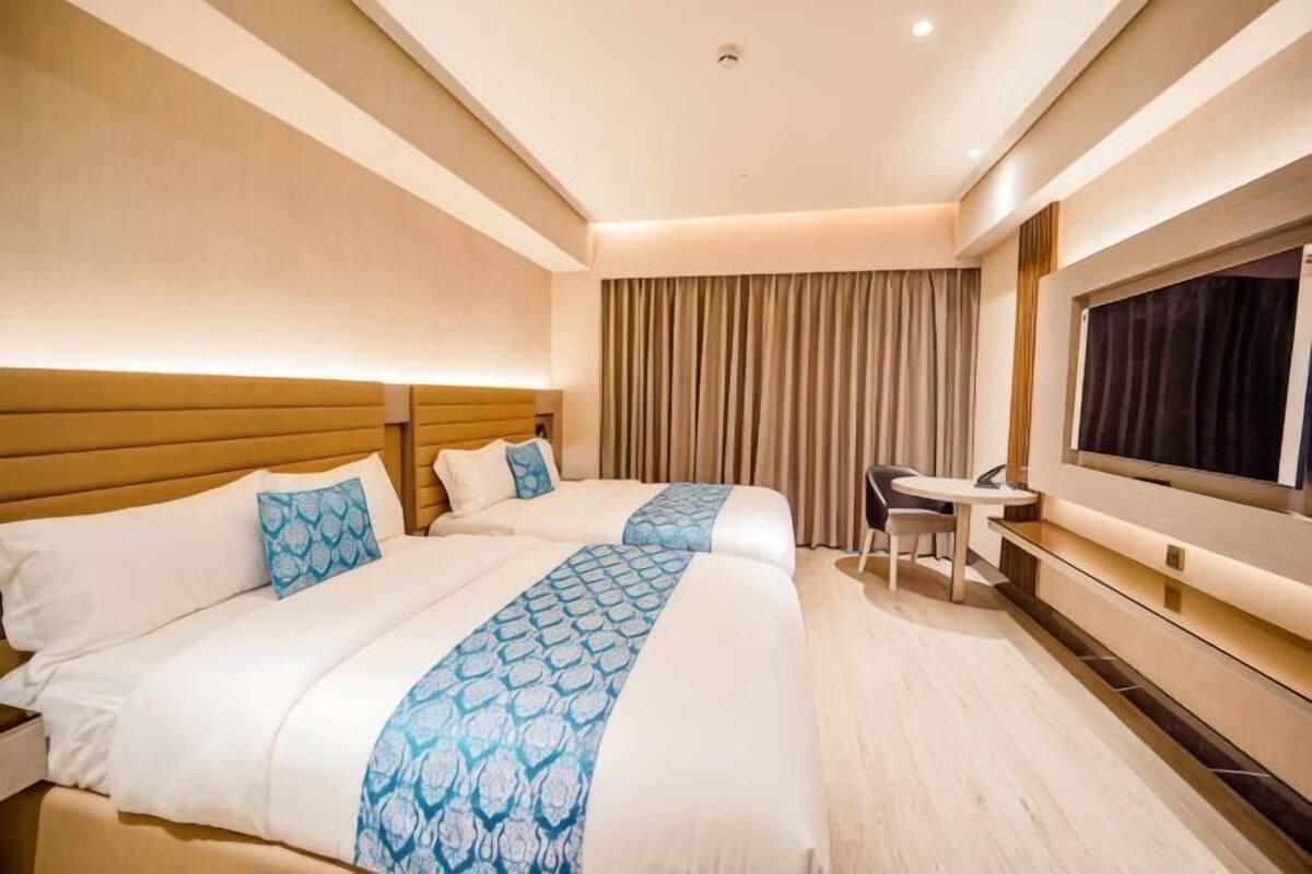 Ein Hotelzimmer mit zwei Betten, einem Schreibtisch, einem Stuhl, einem Fernseher und einem Fenster mit Vorhängen.